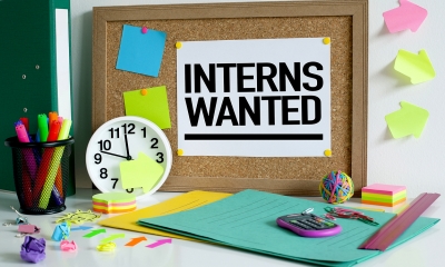 'INTERNS WANTED' sign pinned to cork board with multiple stationary items surrounding