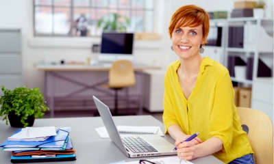 Happy smiling red haired woman in yellow blouse sitting at desk with laptop, eyeglasses and stacks of papers and folders Happy smiling red haired woman in yellow blouse sitting at desk with laptop, eyeglasses and stacks of papers and folders