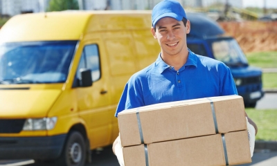 Male courier wearing a blue uniform holding two cardboard boxes in front of a yellow van