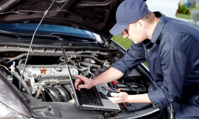 Man working in a garage using his laptop whilst fixing a car