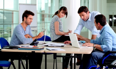 Disabled man sat at a desk using his laptop with a group of employees