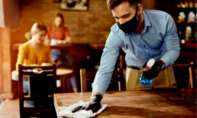 A waiter cleans a cafe table while wearing gloves and a face mask