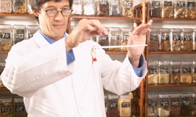 Man with glasses in white lab coat mixing herbs in front of rows of herb jars