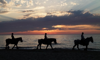 Three people trekking on ponies along a beach with the sunset in background