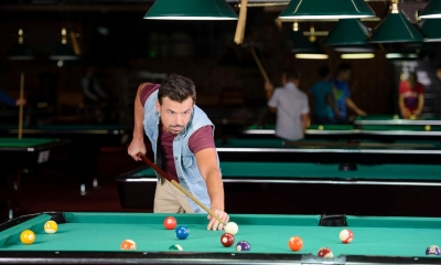 Man in waistcoat playing pool in dark room with peoples and tables in background