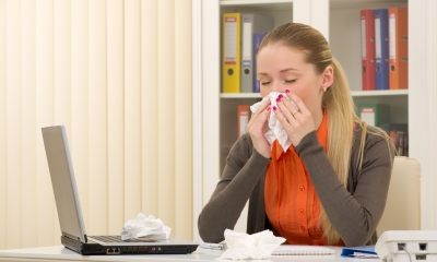 Sickness and sick pay Young woman sits at her desk blowing her nose into a tissue