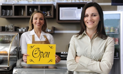 Starting up Two women standing at the counter of their cake shop, one holding a yellow sign with open written on it