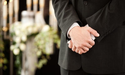 Close up of a funeral director standing in front of some flowers with his hands clasped