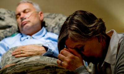 Woman with her head in her hands with an ill man in bed in the background